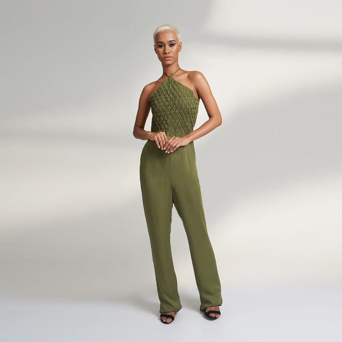 A global small size model wearing a solid olive green jumpsuit, highlighted with hand-stitched smocking on the bodice panel with a drawstring attached to the bodice to tie at the neck. inspired from the vintage window designs.