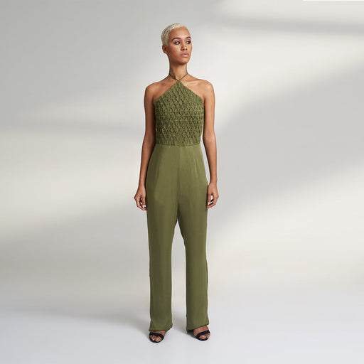 A global small size model wearing a solid olive green jumpsuit, highlighted with hand-stitched smocking on the bodice panel with a drawstring attached to the bodice to tie at the neck. inspired from the vintage window designs.