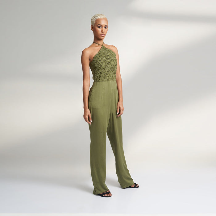 A global small size model wearing a solid olive green jumpsuit, highlighted with hand-stitched smocking on the bodice panel with a drawstring attached to the bodice to tie at the neck. inspired from the vintage window designs.