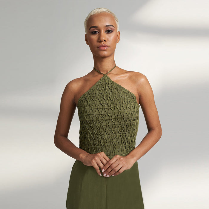 A global small size model wearing a solid olive green jumpsuit, highlighted with hand-stitched smocking on the bodice panel with a drawstring attached to the bodice to tie at the neck. inspired from the vintage window designs.